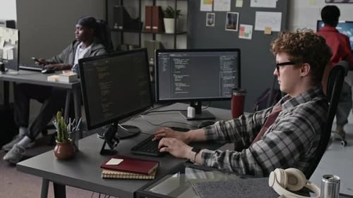 Man Coding on Dual Monitors with Multiethnic Team in Tech Office