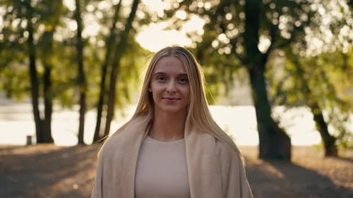 A beautiful peaceful girl covers herself with a blanket on the river bank near park and squints