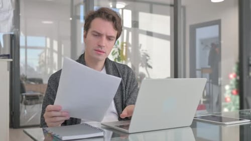 Man Works on Laptop and Reviews Documents