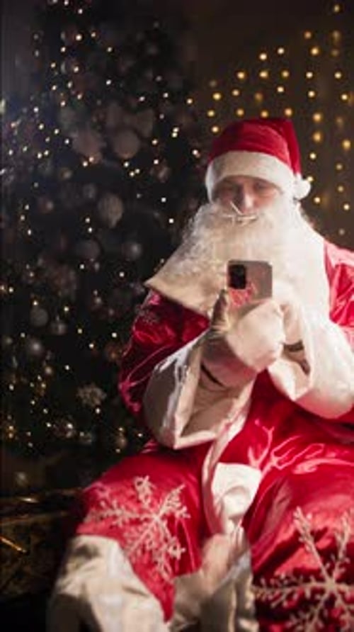Man in Santa Costume with Smartphone in Hands Reading Messages Sitting Against Christmas Tree and