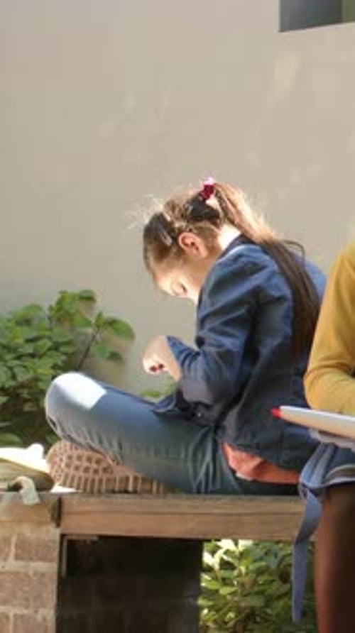 Vertical video: Sunlight dappling youth in denim fixing red item on bench studying with notebook