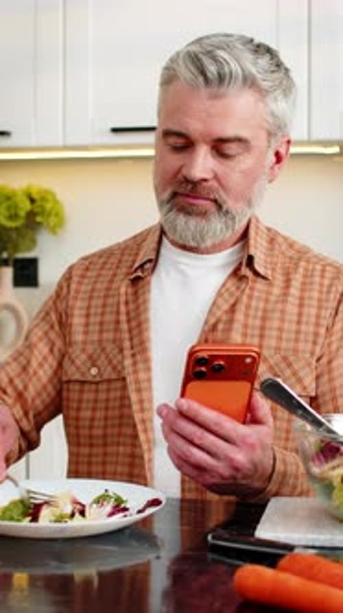 Middleaged Man Eating Healthy Diet Salad Using Smartphone for Diet Tracking in Home Kitchen Indoors