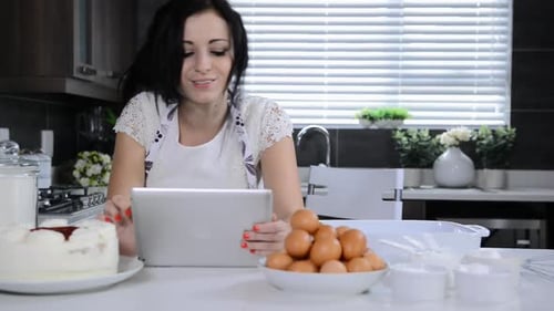 Cheerful brunette woman uses tablet computer while baking cupcakes in kitchen