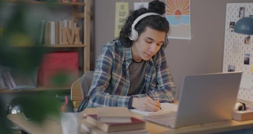 Teenage Boy Doing Homework Using Laptop and Writing in Notebook Using Headphones at Table at Home