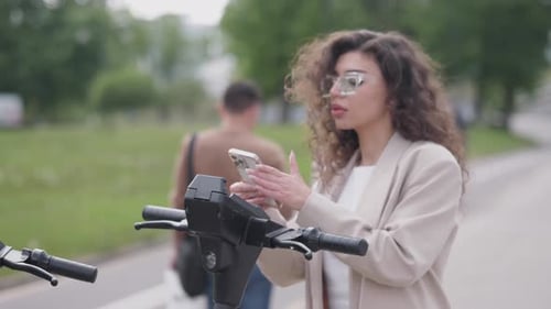 A Girl Using an Application on a Gadget Tries to Rent an Electric City Scooter in Summer
