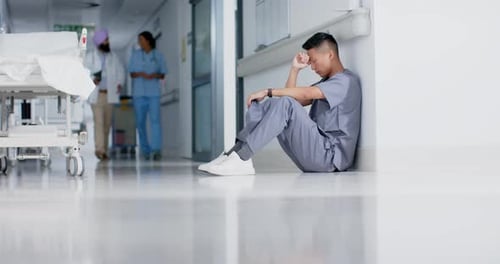 Stressed asian male surgeon sitting and leaning on wall in hospital in slow motion