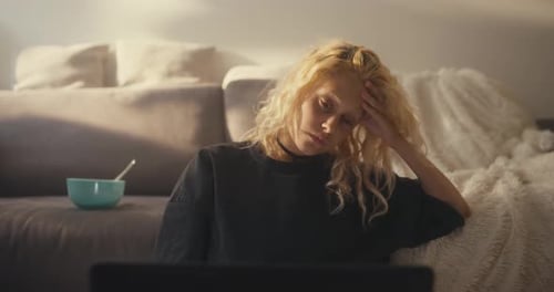 A Tired Blonde Girl in a Gray Tshirt Leans on Her Hand and Looks Sadly at the Laptop Screen Near the