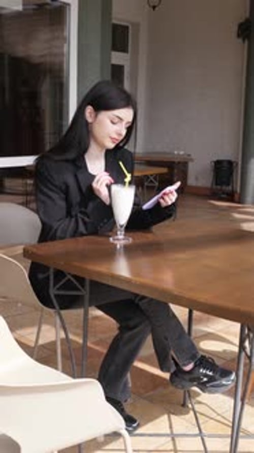 86 Una niña usa un teléfono inteligente y bebe un cóctel mientras está sentada en una mesa en la calle.