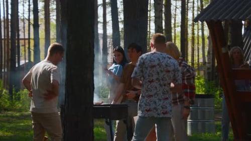 Friends are standing at barbecue and cooking meat in forest