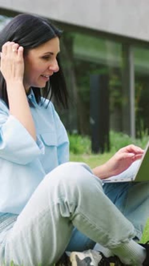 Selbstbewusste junge Frau in Freizeitkleidung arbeitet am Laptop, während sie auf grünem Gras im Stadtpark sitzt