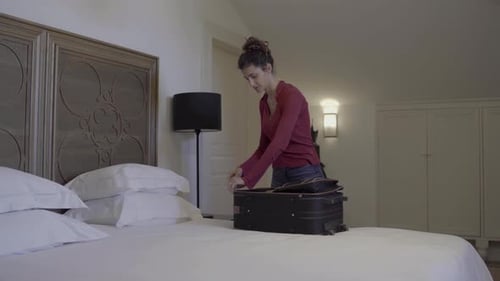 A young curly-haired woman packing her suitcase on a bed hotel bed and leaving.