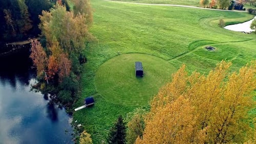 Aerial shot of grass field with barrel sauna next to natural lake during daytime. Idyllic landscape