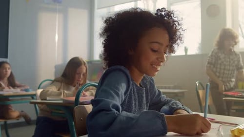 Positive african american schoolgirl smiling and writing notes during elementary school lesson