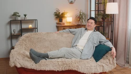 Hombre chino de mediana edad feliz y despreocupado sentado cómodamente en el sofá pensando en la sala de estar de su casa