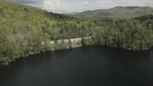 Drone shot flying backwards over water, with a red car and 2 people on the shore, revealing a forest