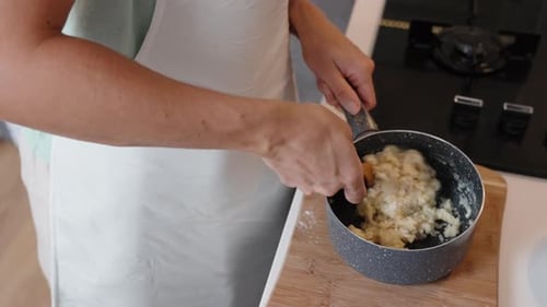 The woman is stirring choux pastry in a saucepan on a wooden cutting board.