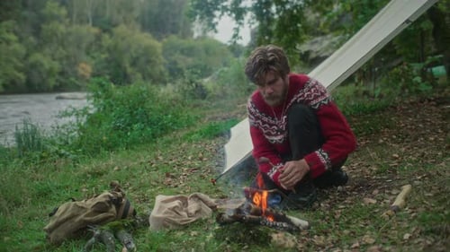Man Cooking Food over Fire beside River and Tent during Solo Camping Trip