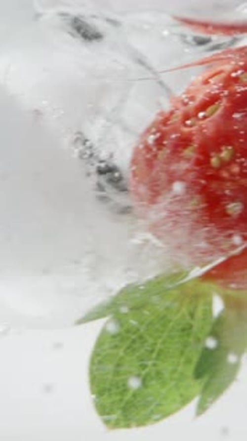 Strawberries and ice cubes in a soda glass rotate and bubbles rise up, macro on a black background.