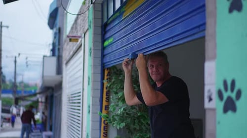 Man closing storefront garage in city sidewalk. Person slides storefront in street