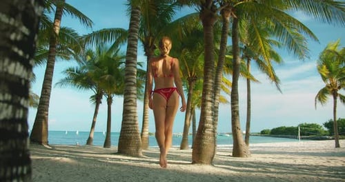 Woman walking barefoot in swimsuit between palm trees on a sunny tropical beach