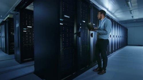 Bearded IT Specialist in Glasses is Working on Laptop in Data Center while Standing Before Server R