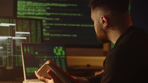 Closeup View of a Young Man Sitting in His Room in Front of Several Monitors and Reading a Book A