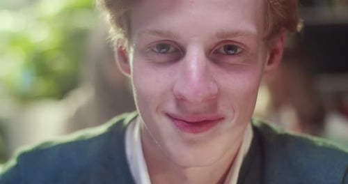 Portrait of Young Cheerful Red Haired Caucasian Male Student Sitting at Table in Library and Smiling