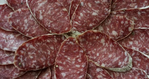 Macro shot of a salami on a wooden cutting board. One hand takes the knife and slices of slices to