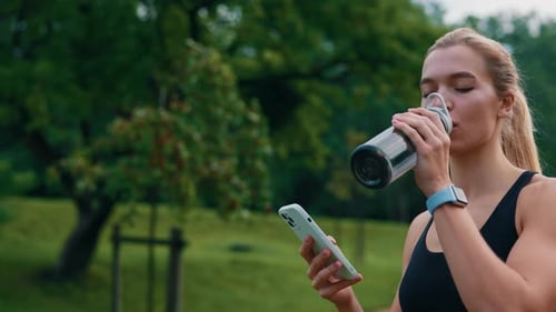 Close Up of the Sporty Woman Walking in the Park After Running and Using Smartphone