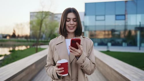 Graceful Caucasian Woman Walking Along the Local Lake Chatting in Mobile Phone with Coffee in Hand