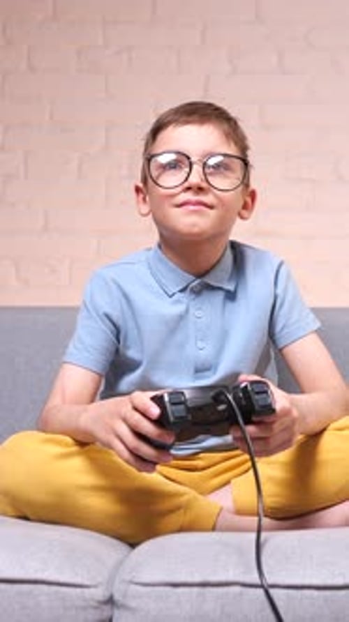 Vertikales Videoporträt eines männlichen Kindes mit Gamepad, das auf dem Sofa sitzt