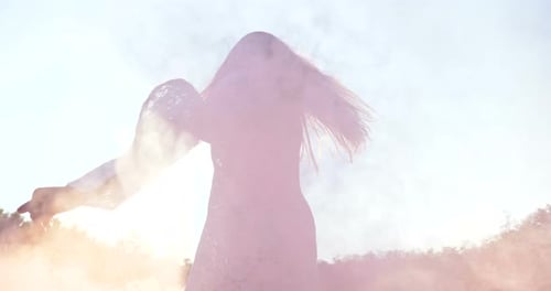 On a sunny day, a woman walks across the field with a colored smoke flare in a white dress against
