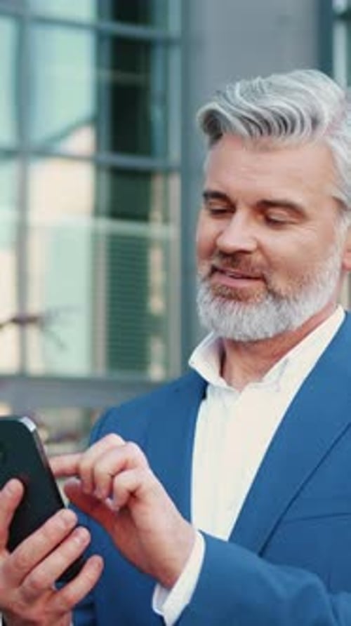 Senior Greyhaired Bearded Businessman in a Formal Suit Standing Outdoors Using Smartphone to Browse