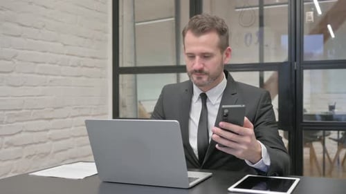 Man in Suit Using Phone and Laptop at Desk