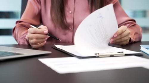 Close-up of female hand signing documents at desk in office. Businesswoman carefully reviewing