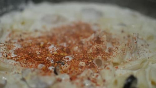 Close Up View Of Spices Being Sprinkled Onto Bubbling Macaroni Pasta In Cream Sauce. Low Angle, Slow
