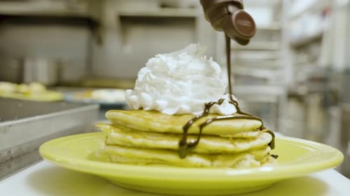 Close-up of sauce being drizzled over a stack of pancakes with whipped cream on top