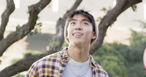 Asian man noticing overhead, tilting head upward in park by tree trunk, facing camera and grinning