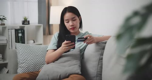 Young woman uses phone and credit card to shop online on sofa at home.