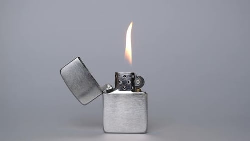Un briquet en métal gris brûle à la lumière sur un fond gris, allumant un feu avec un briquet sur fond gris