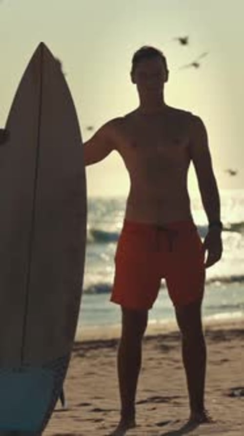 Surfer Holding Surfboard on Beach at Sunset with Birds Flying