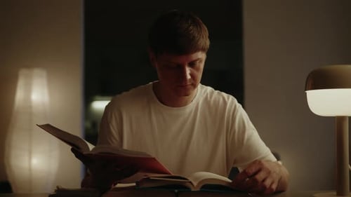Male Student Concentrating Reading Two Paper Books Sitting at a Table in Dimly Lit Home Interior