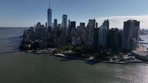 An aerial view of lower Manhattan and New York Harbor on a sunny day with blue skies. The camera fac