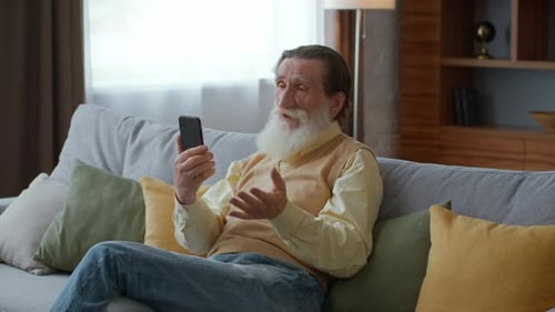 Bearded Senior Man Video Chatting with Friends or Family Discussing World News Sitting on Sofa at