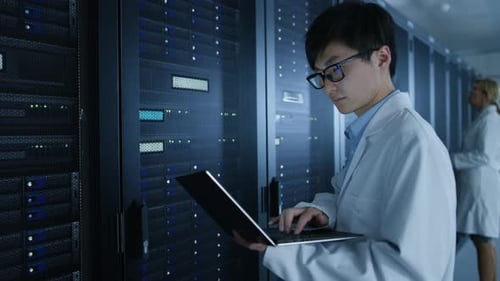 Im Rechenzentrum: Männliche und weibliche IT-Spezialisten in weißen Kitteln arbeiten mit Server-Racks, nutzen Lapt
