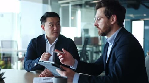Diverse Partners Discussing Financial Project on Tablet While Sitting By Desktop