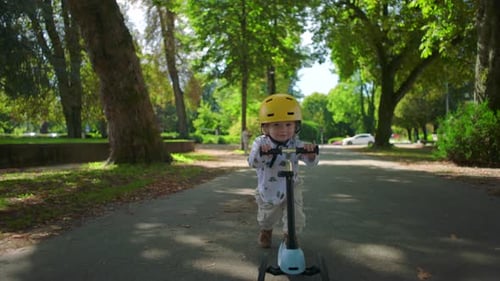 Small toddler wearing yellow helmet riding scooter along tree-lined park path, moving with