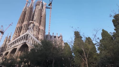 Iconic Sagrada Familia Cathedral Surrounded by Trees Against a Clear Blue Sky. Pan Left