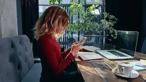 Happy Young Woman Sits with Phone and Computer at the Table
