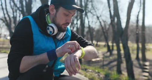 While Running in the Park Young Man with Beard Squats Down and Looks at How Far He Has Run on Watch